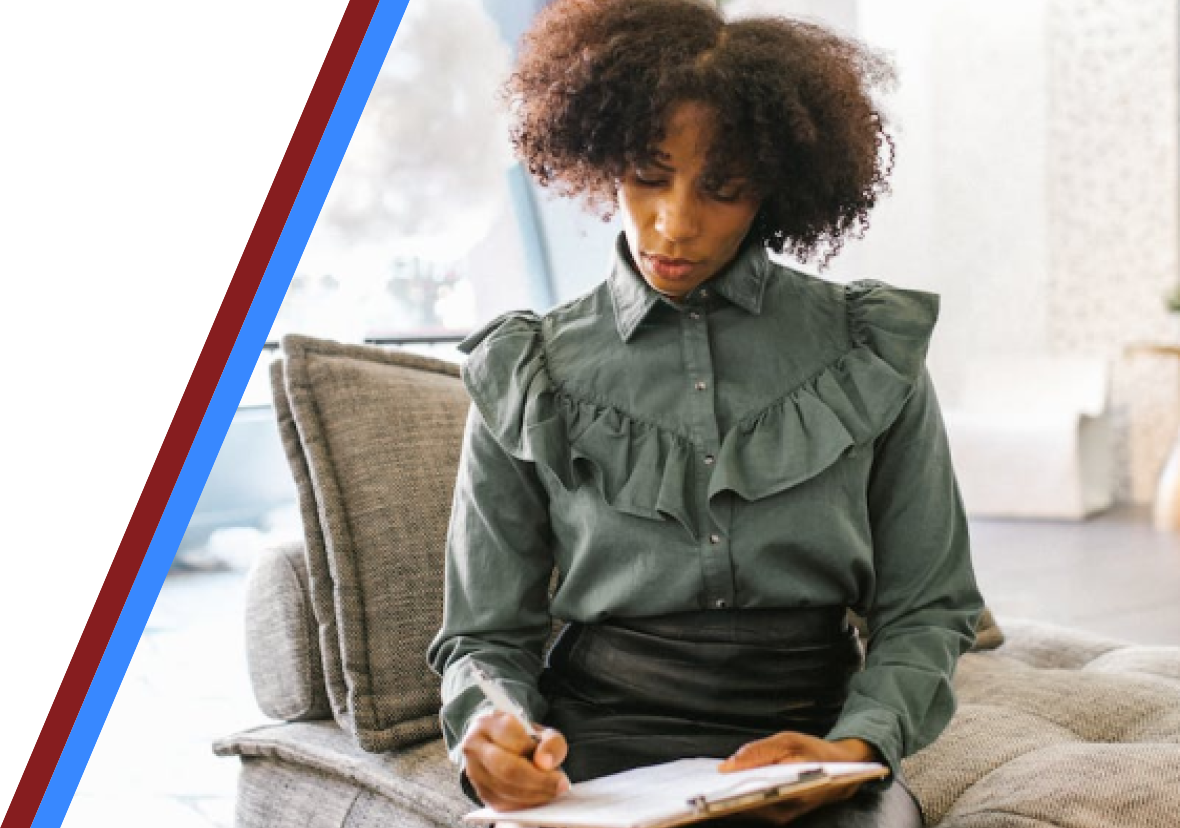 A black woman with afro hair in office clothes sitting and writing on a clipboard